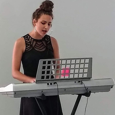 girl in black dress standing in front of white wall while playing the keyboard